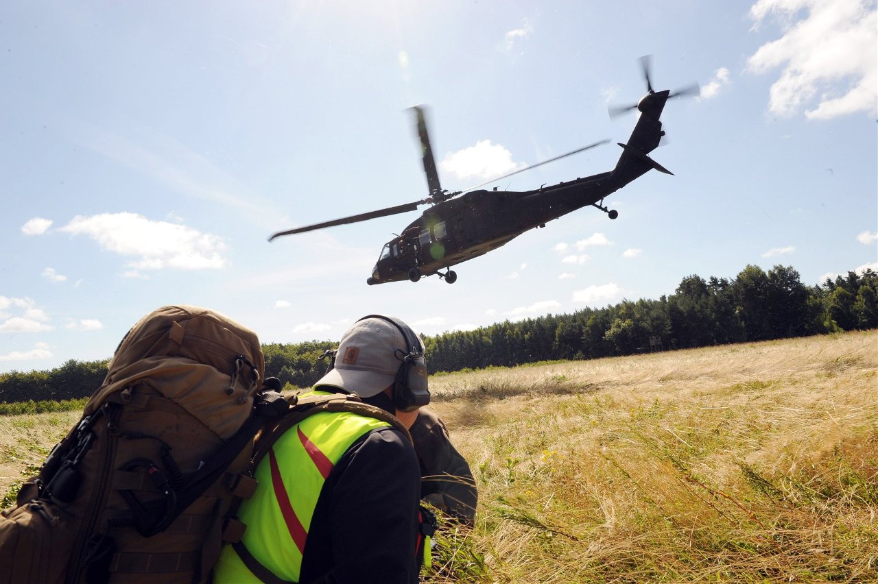 Śmigłowiec Black Hawk nisko nad polaną, na pierwszym planie żołnierz w kamizelce odblaskowej z plecakiem