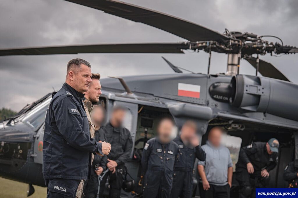 Policjant w mundurze prowadzi odprawę przed lotem, w tle policyjny śmigłowiec S-70i Black Hawk