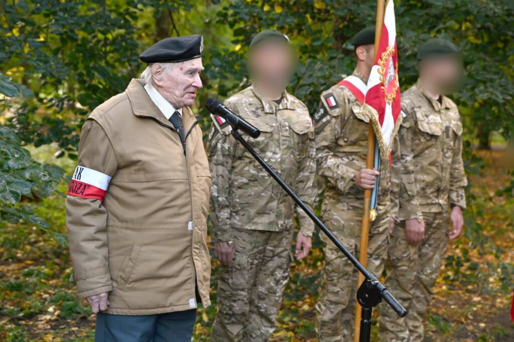 Starszy mężczyzna w berecie i z opaską Armii Krajowej przemawia przy mikrofonie, za nim poczet sztandarowy w mundurach polowych