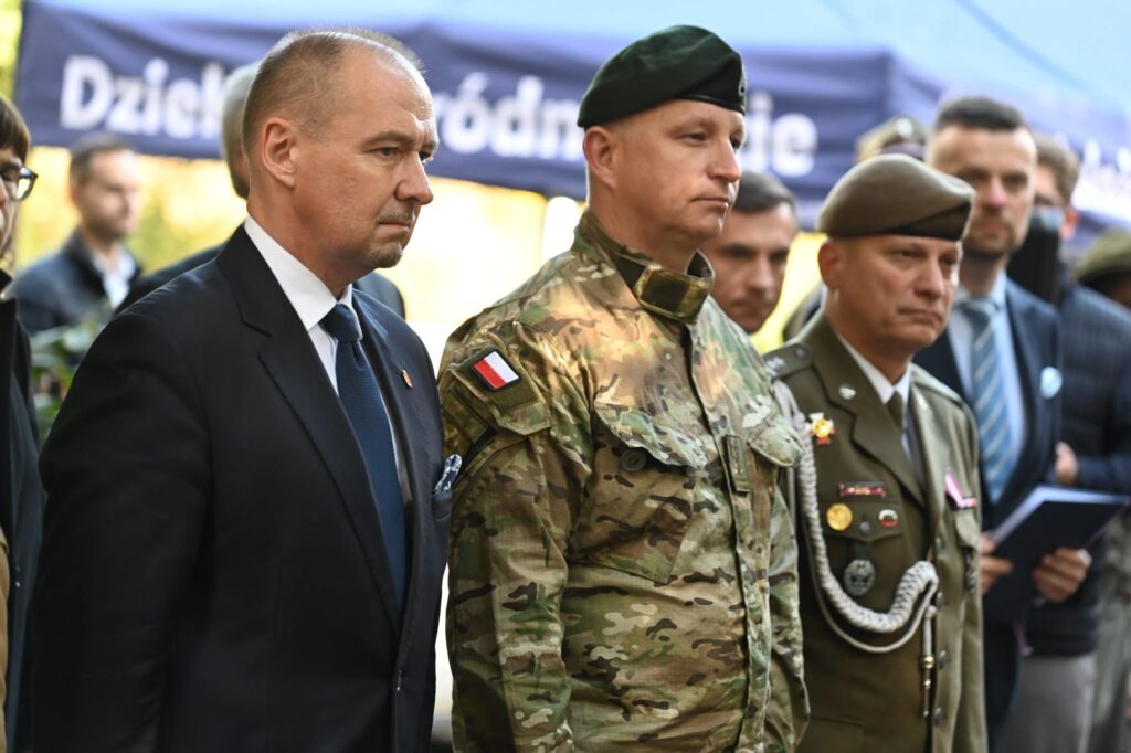 Mężczyzna w garniturze oraz oficerowie w mundurze polowym i wyjściowym stoją w szeregu, w tle baner dzielnicy Śródmieście