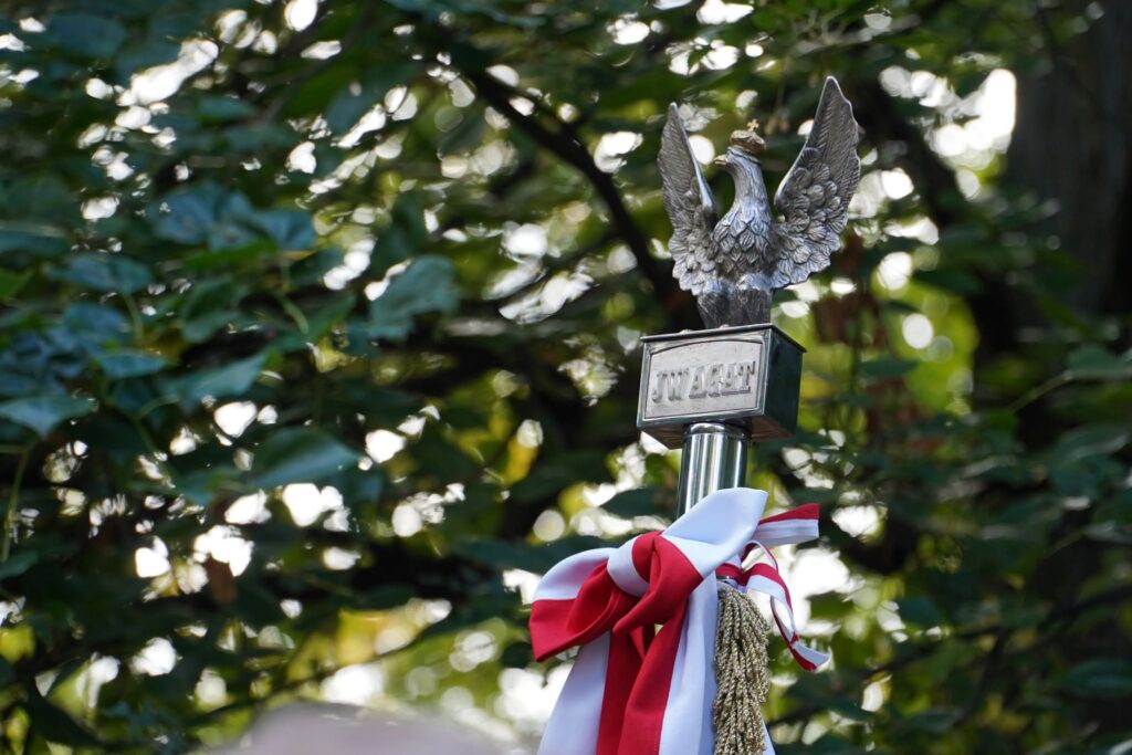 Głowica sztandaru z orłem i tabliczką, przewiązana biało-czerwoną wstęgą, na tle drzew
