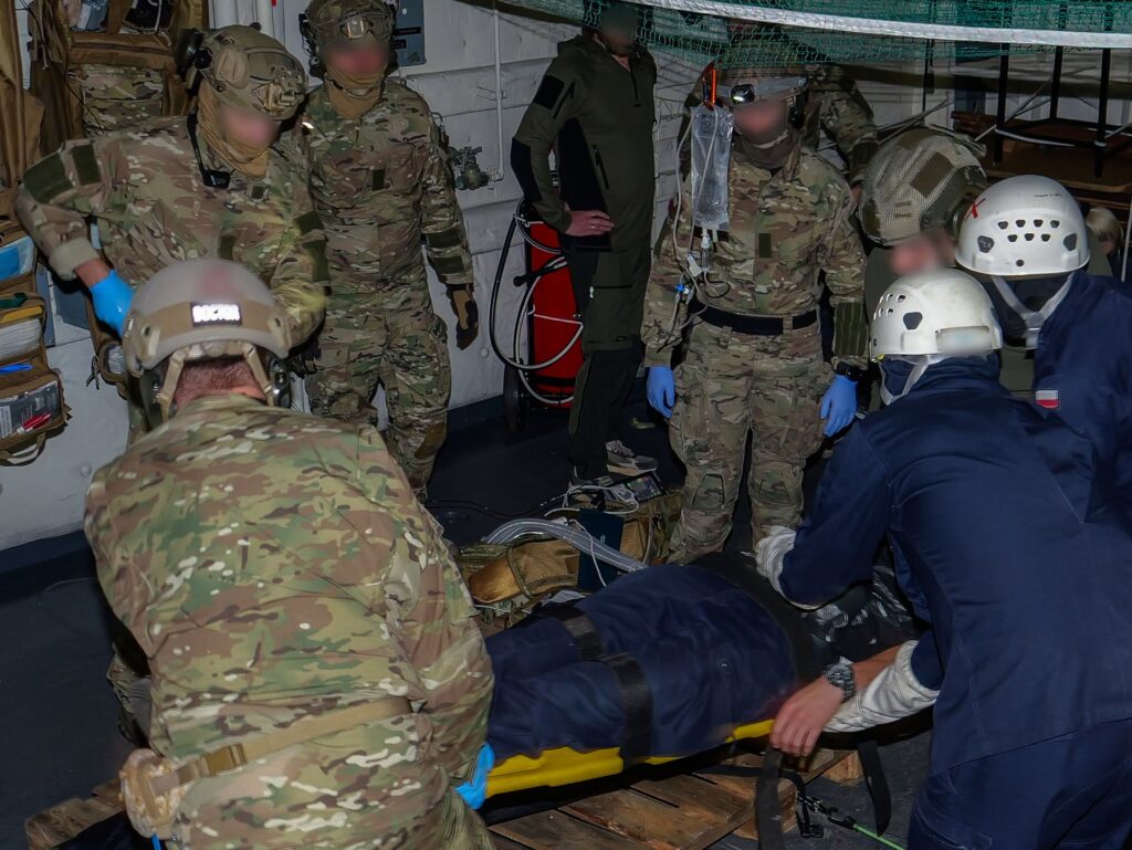 Przenoszenie osoby na noszach — po jednej stronie żołnierze w mundurach polowych, po drugiej ratownicy w białych hełmach