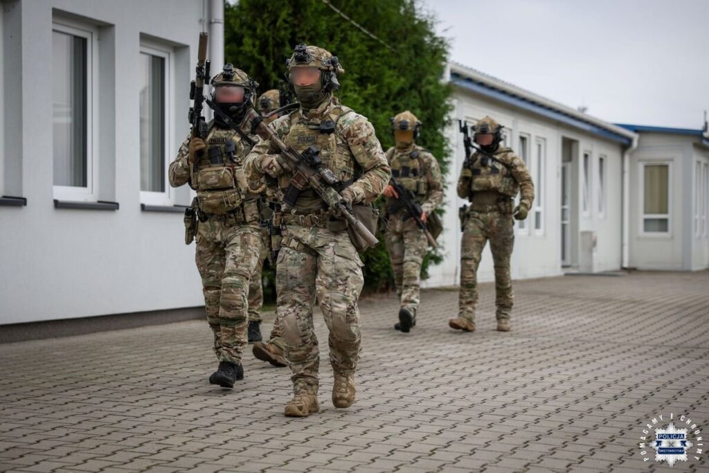Czterech funkcjonariuszy w umundurowaniu taktycznym przemieszcza się kolumną wzdłuż budynku