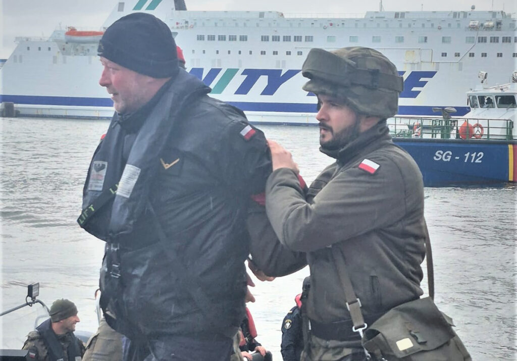 Umundurowany żołnierz prowadzi mężczyznę po nabrzeżu, w tle jednostka pływająca SG-112 i prom